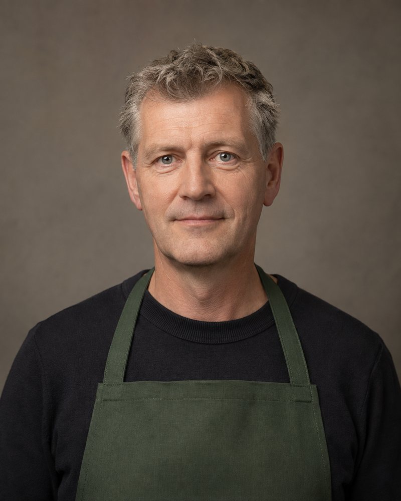 Portret van een man van ongeveer 50 met kort grijs haar en een donkergroene schort